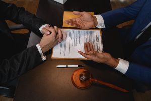 A contract document sits on a dark wood table in an office setting. Two people in tailored suits are discussing the contract agreement at the table. A gavel and fancy pen also sit on the table. This business photo is perfect for projects related to law, business agreements, or finance