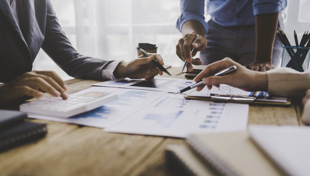 Three people are gathered around a wooden table reviewing charts and graphs. Source: Envato.com
